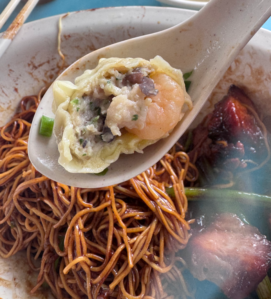 A Chef Kang wanton cut open on a spoon, showing a whole prawn and minced pork filling, above the mixed wanton noodles