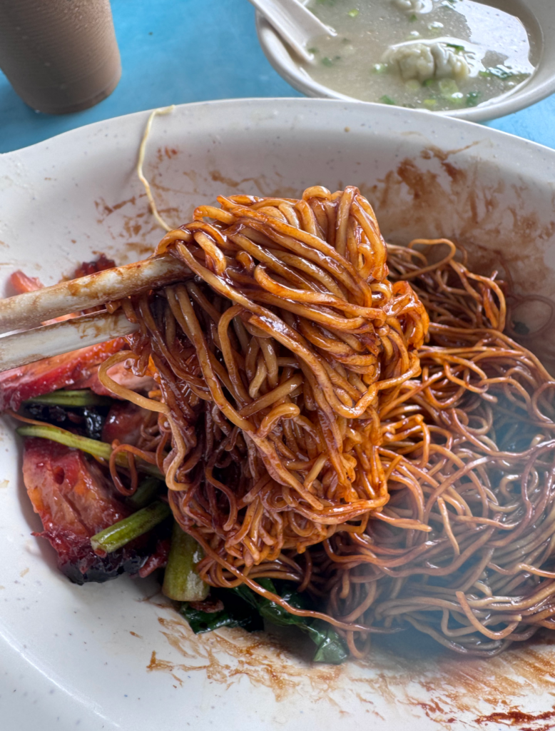 Chef Kang wanton mee noodles lifted with chopsticks after mixing, glossy and coated in black sauce, chilli and oil