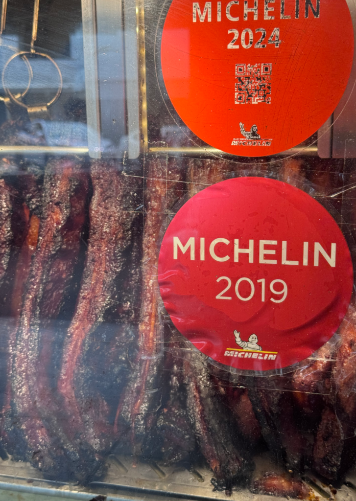 Michelin 2019 and 2024 stickers on the glass display of hanging charcoal-roasted char siew at Chef Kang’s Noodle House