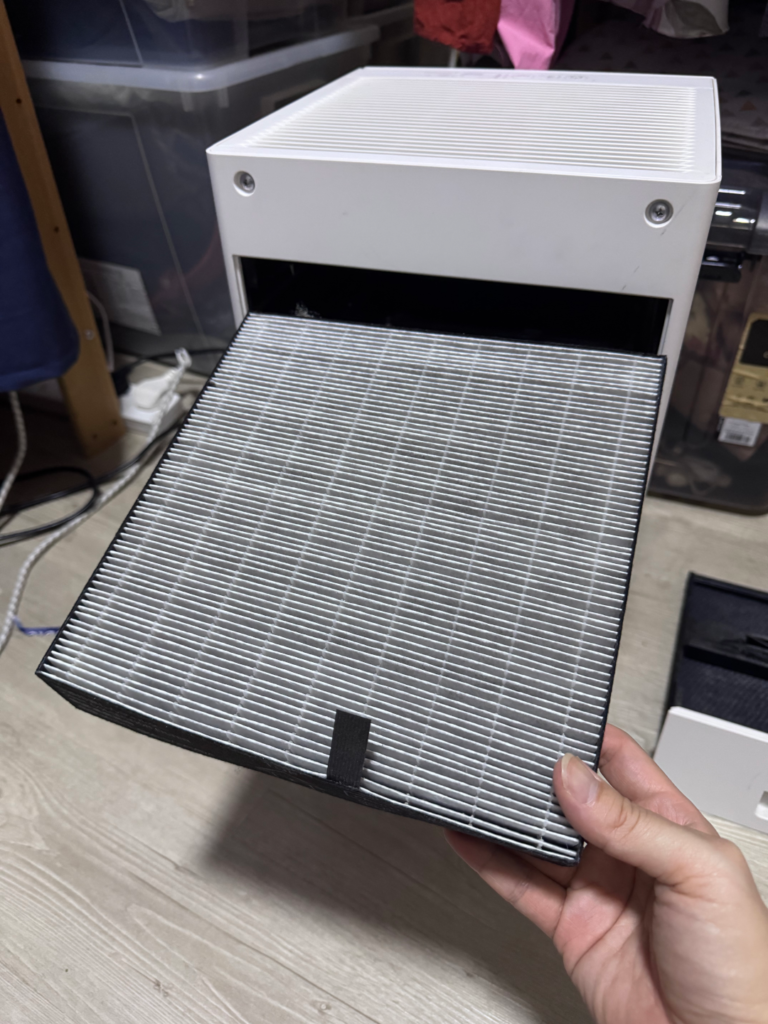 A hand holding the flat, pleated electrostatic HEPA filter from a Daikin MC30YVM7 air purifier, lightly greyed with dust after use.